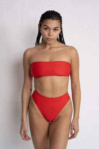 A woman standing in front of a white wall with her arms by her side wearing bright red high waisted bikini bottoms with a matching bright red strapless bandeau bikini top. 