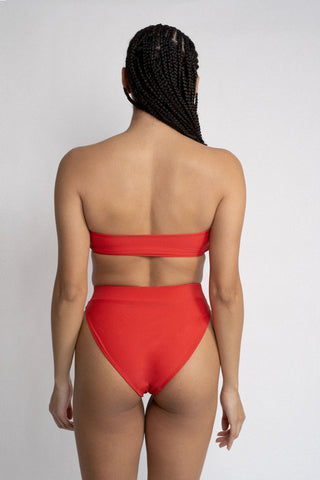 The back of a woman standing in front of a white wall wearing bright red high waisted bikini bottoms with a matching bright red strapless bandeau top.
