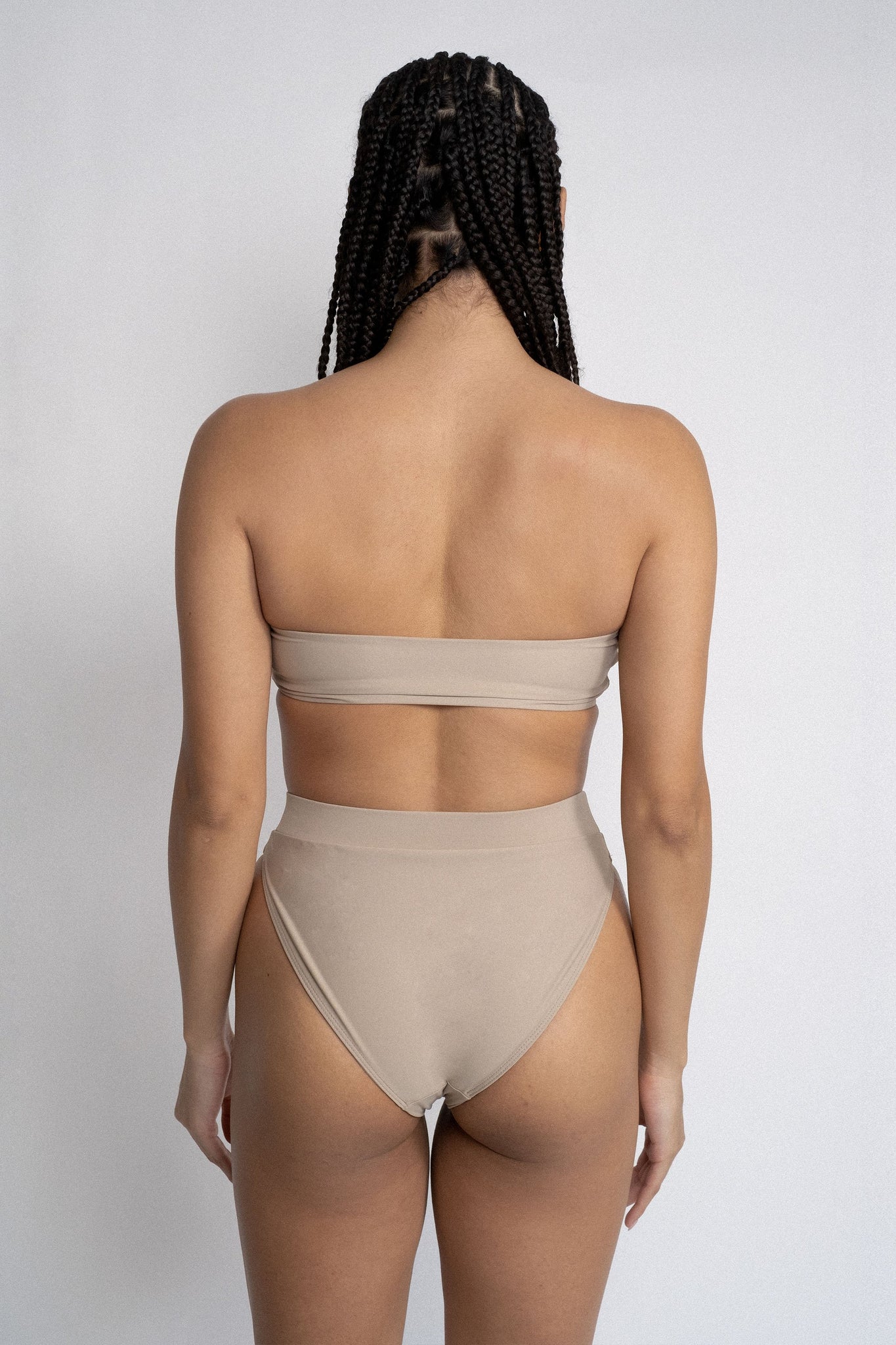 The back of a woman standing in front of a white wall wearing nude high waisted bikini bottoms with a matching nude bandeau strapless bikini top.