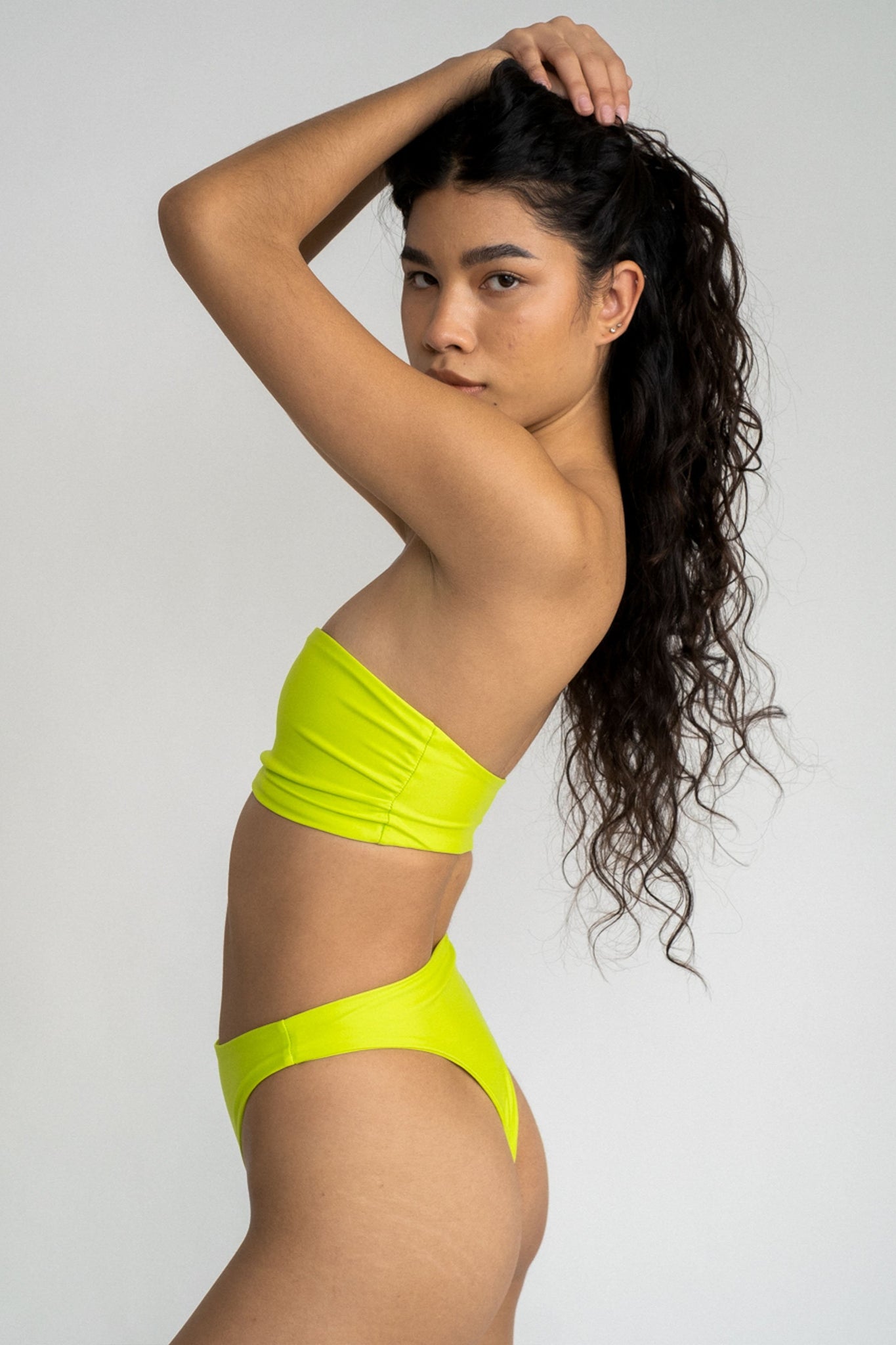 A woman standing to the side with her arms above her head wearing bright neon green high cut bikini bottoms with a matching bright green strapless bandeau bikini top.