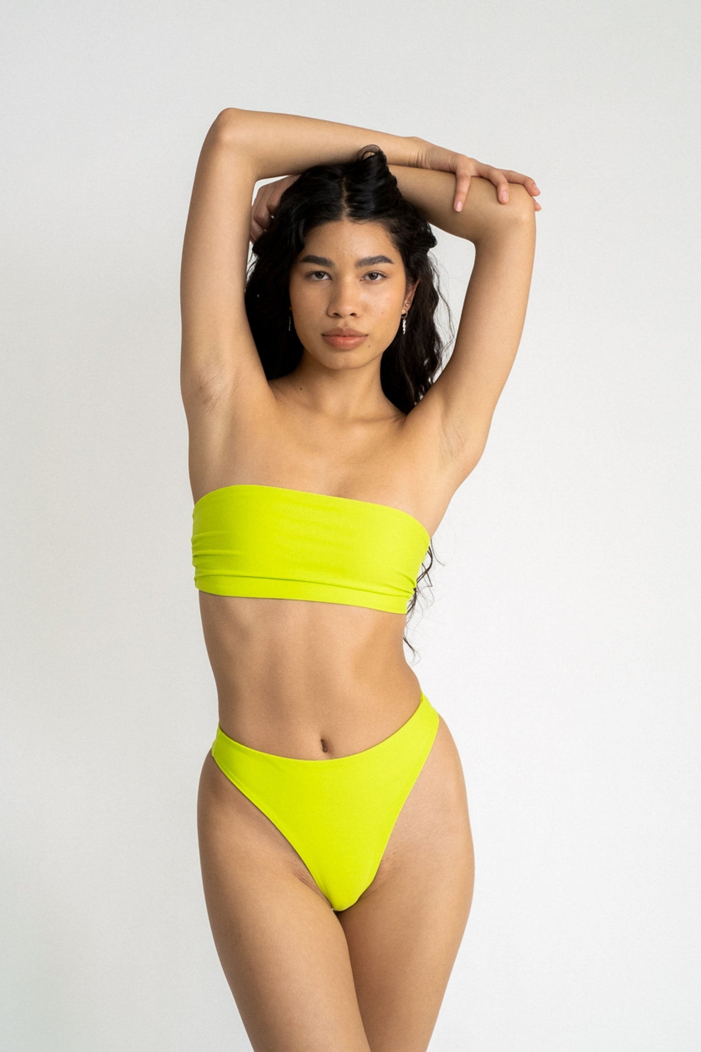 A woman standing with her arms folded above her head wearing neon green high cut bikini bottoms with a matching neon green strapless bandeau bikini top.