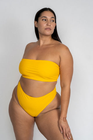 A woman standing looking to the side wearing yellow high cut bikini bottoms with a matching yellow strapless bandeau bikini top.