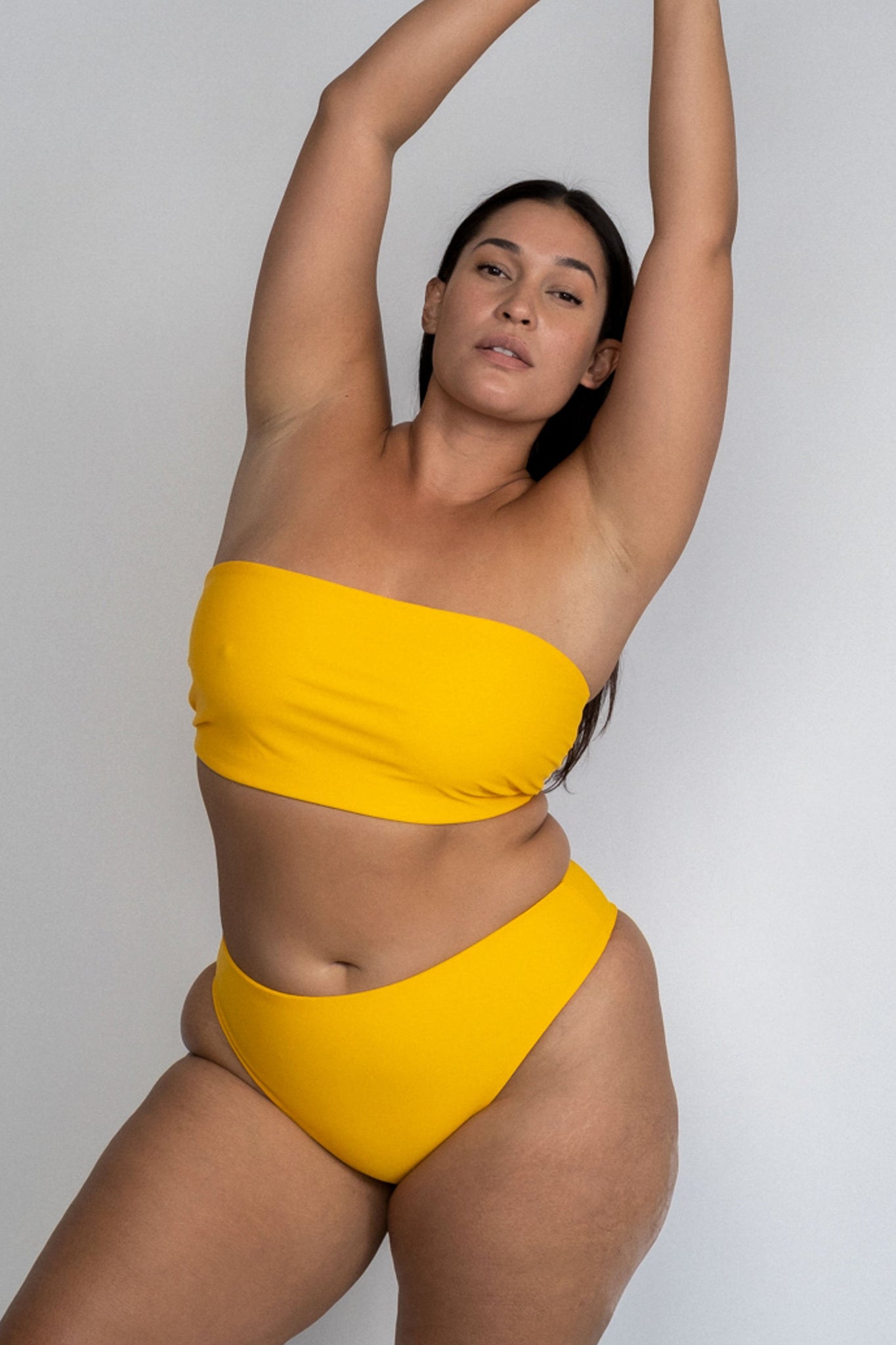 A woman leaning to the side with her arms stretched above her head wearing yellow high cut bikini bottoms with a matching yellow strapless bandeau bikini top.