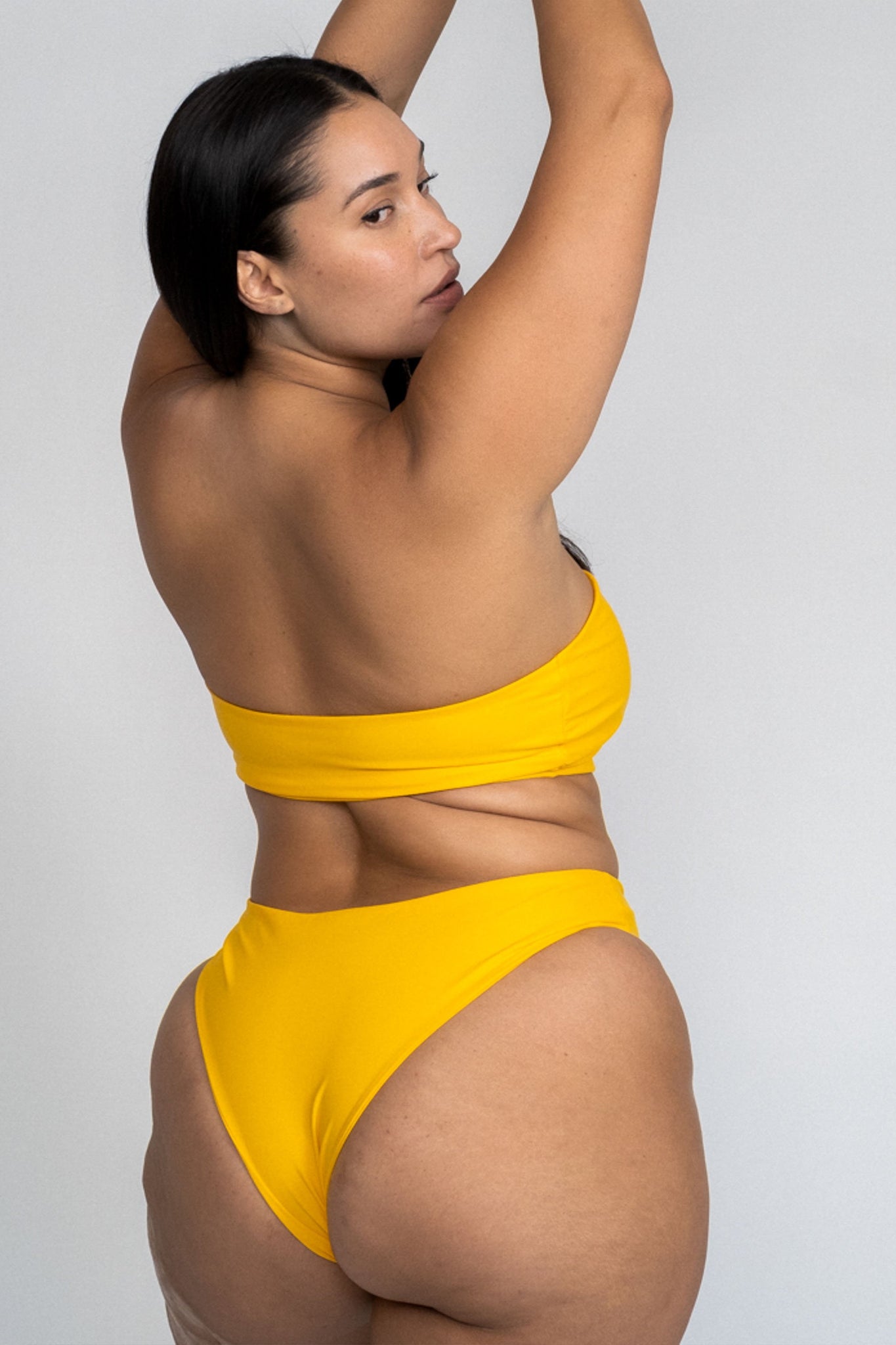 The back of a woman standing looking over her shoulder wearing yellow high cut bikini bottoms with a matching yellow strapless bandeau bikini top.
