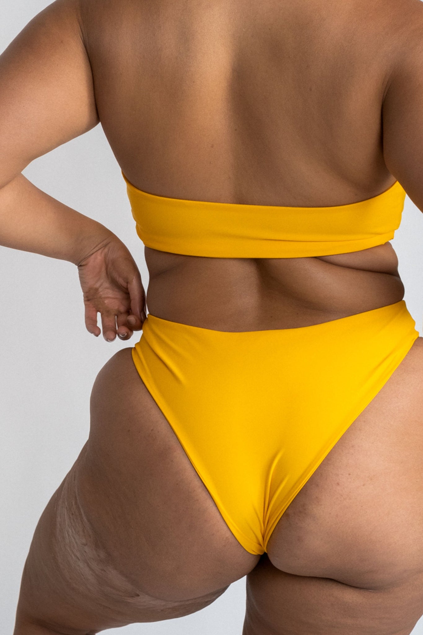 A close up of the back of a woman standing with her hands on her hips wearing yellow high cut bikini bottoms with a matching yellow strapless bandeau bikini top.