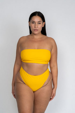 A woman standing with her arms by her side wearing dark yellow high waisted bikini bottoms with a matching dark yellow strapless bandeau bikini top.