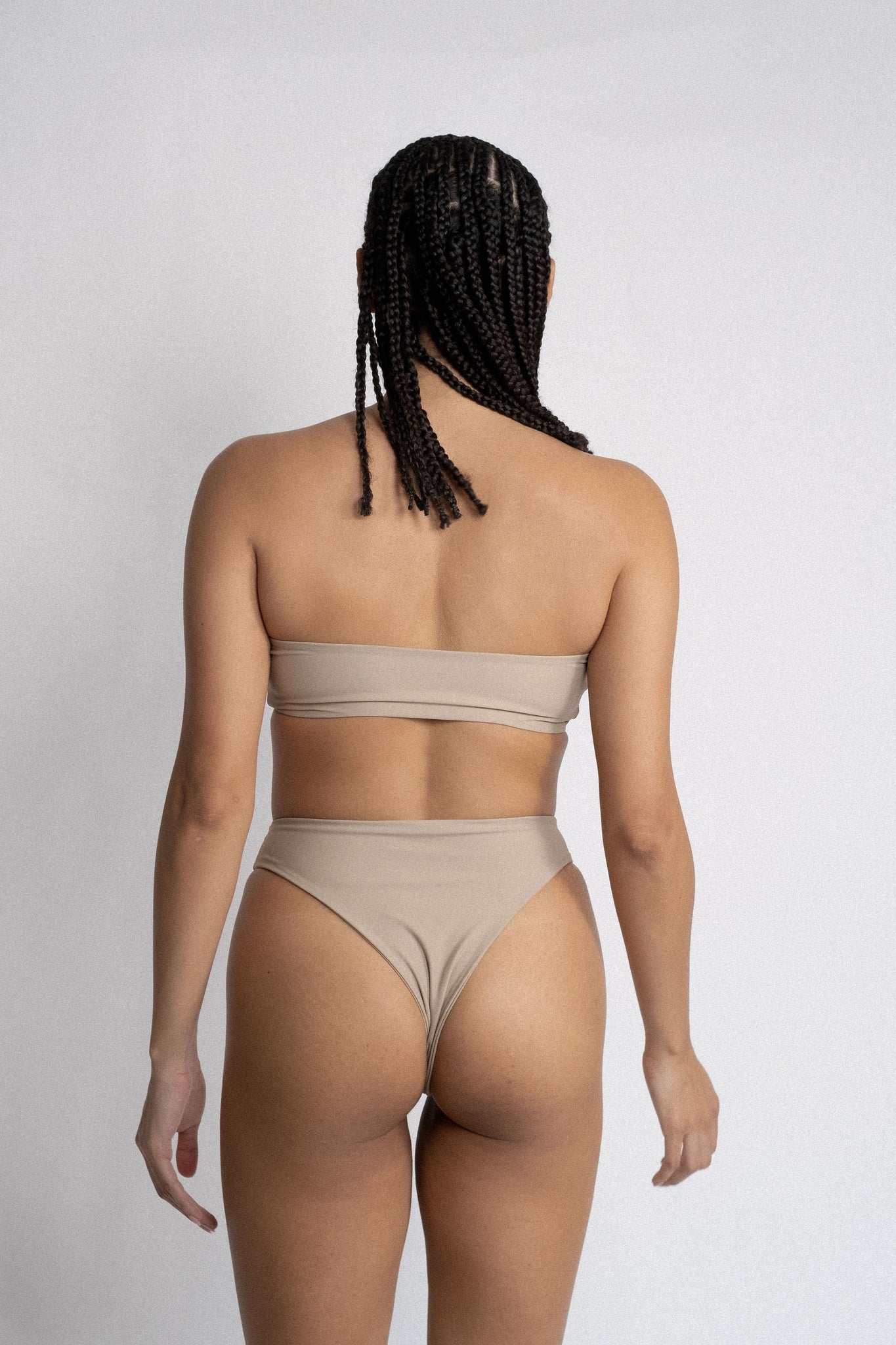 The back of a woman standing in front of a white wall wearing nude high waisted bikini bottoms with a matching nude strapless bandeau bikini top.