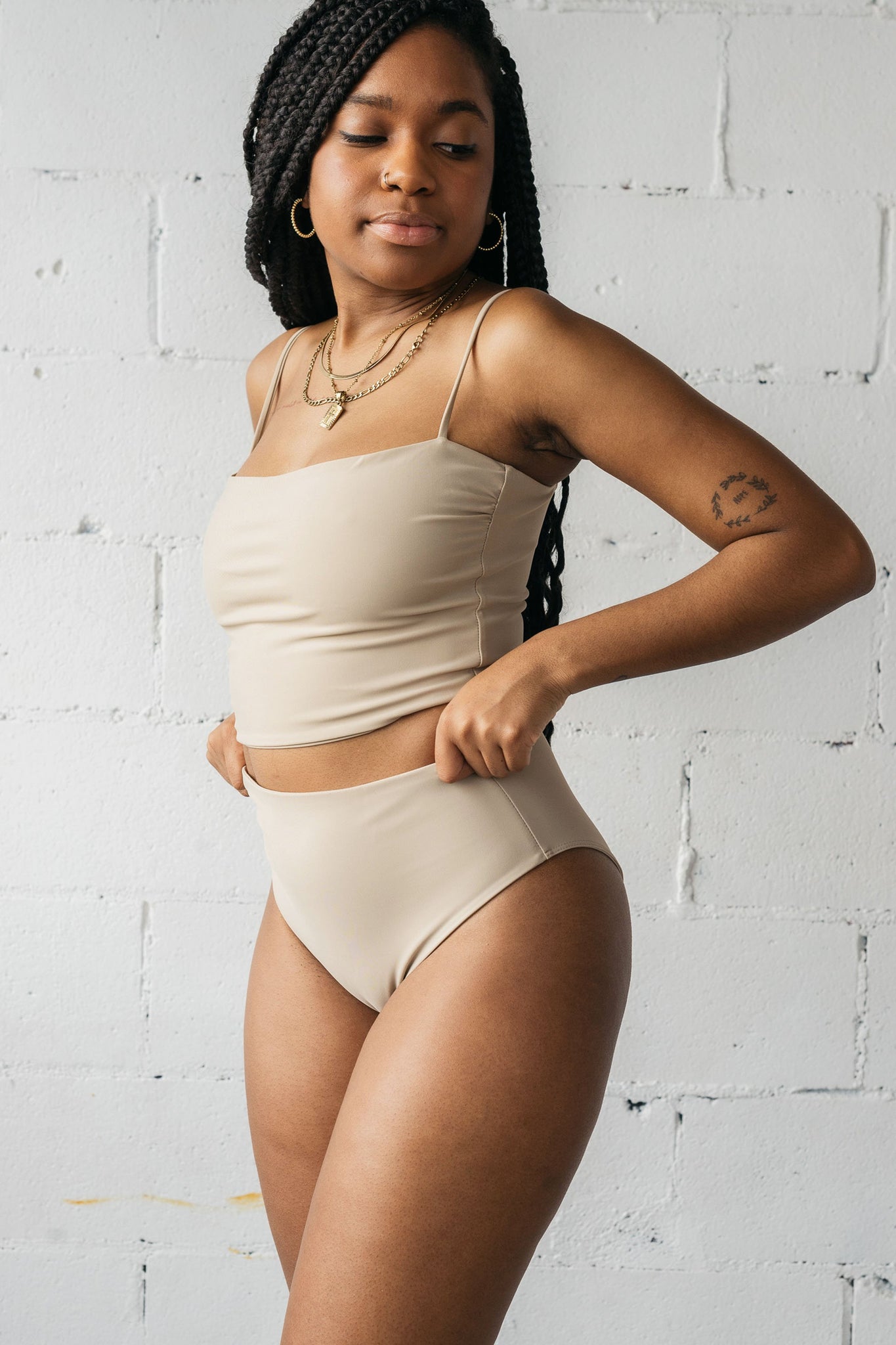 A woman standing in front of a white wall and looking down with her hands on her hips wearing nude high waisted bikini bottoms and a matching nude tankini top.