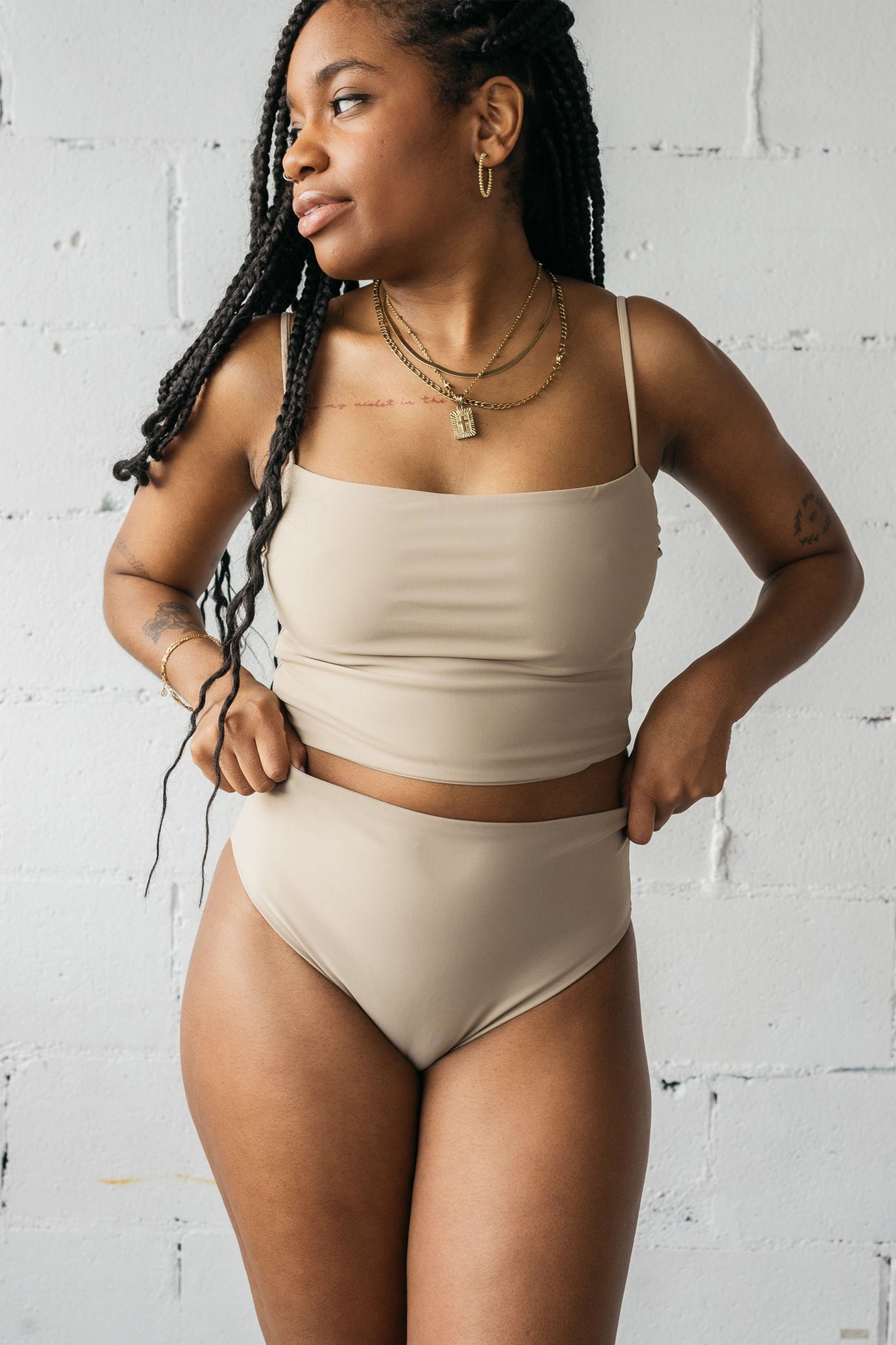 A woman standing looking to the side wearing nude high-waisted bikini bottoms and a matching nude tankini bikini top.