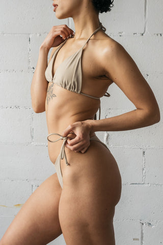 A close up of a woman to the side wearing nude adjustable string bikini bottoms with a matching nude triangle string bikini top.