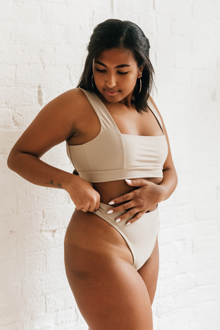 A woman looking down with one arm on her stomach wearing beige high waisted bikini bottoms and a matching beige bikini top with a square neckline.