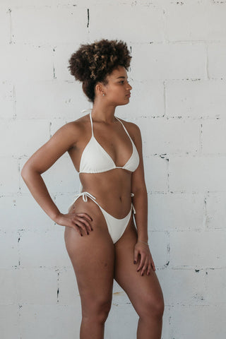 A woman standing with one hand on her hip looking to the side wearing white triangle string bikini bottoms with a matching white triangle string bikini top.