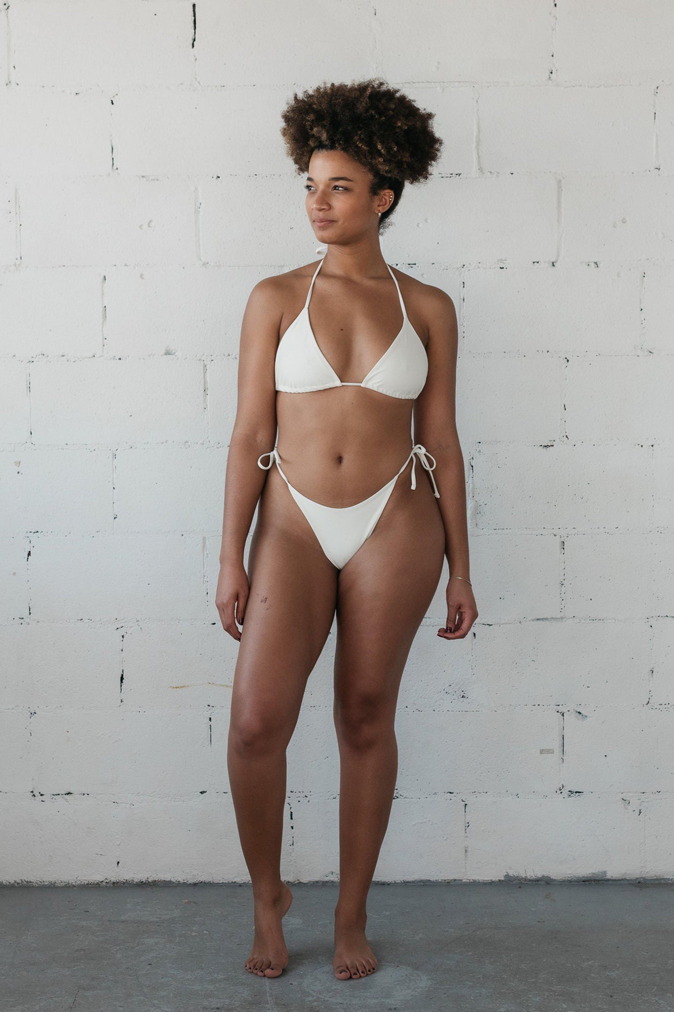 A woman standing with her arms relaxed by her sides wearing white triangle string bikini bottoms with a matching white triangle string bikini top.