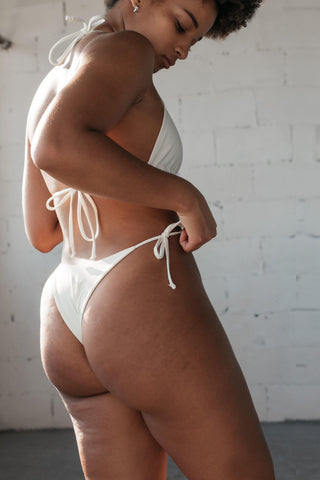 The back of a woman looking down with one hand adjusting her bathing suit wearing white triangle string bikini bottoms with a matching white triangle string bikini top.