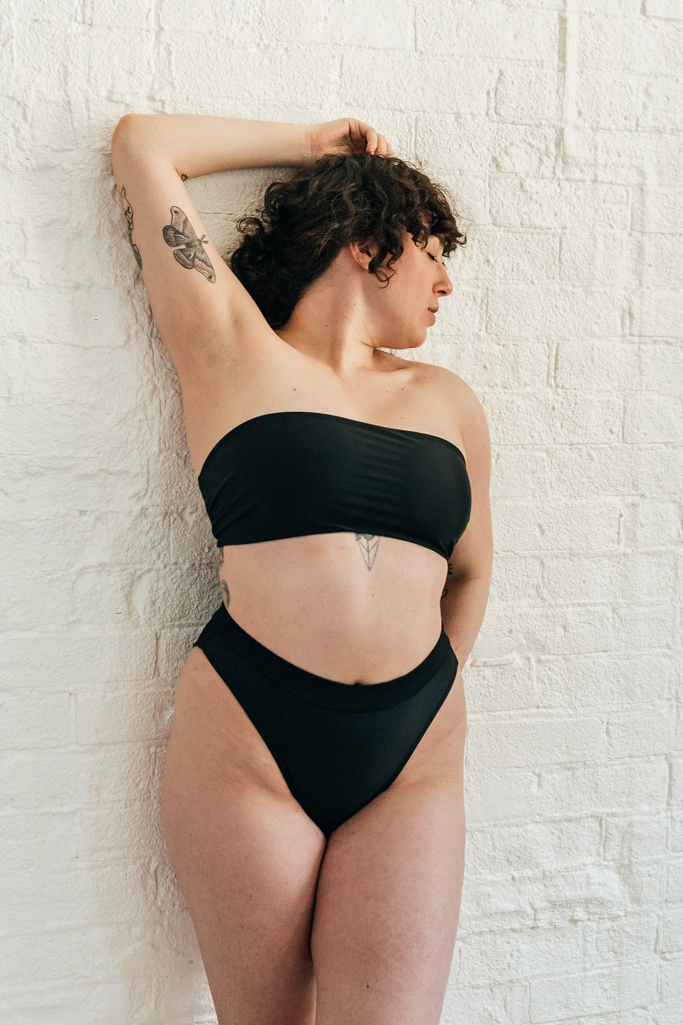 A woman leaning against a white wall looking to the side wearing black high waisted bikini bottoms with a matching black bandeau bikini top.