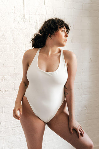 A woman standing in front of a white brick wall wearing a white high cut one-piece swimsuit.