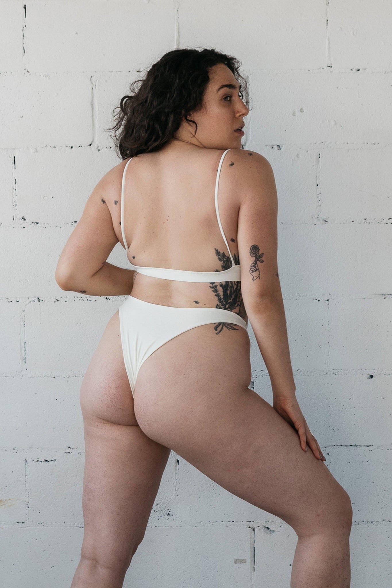 The back of a woman standing against a wall wearing a minimalist white bikini 
