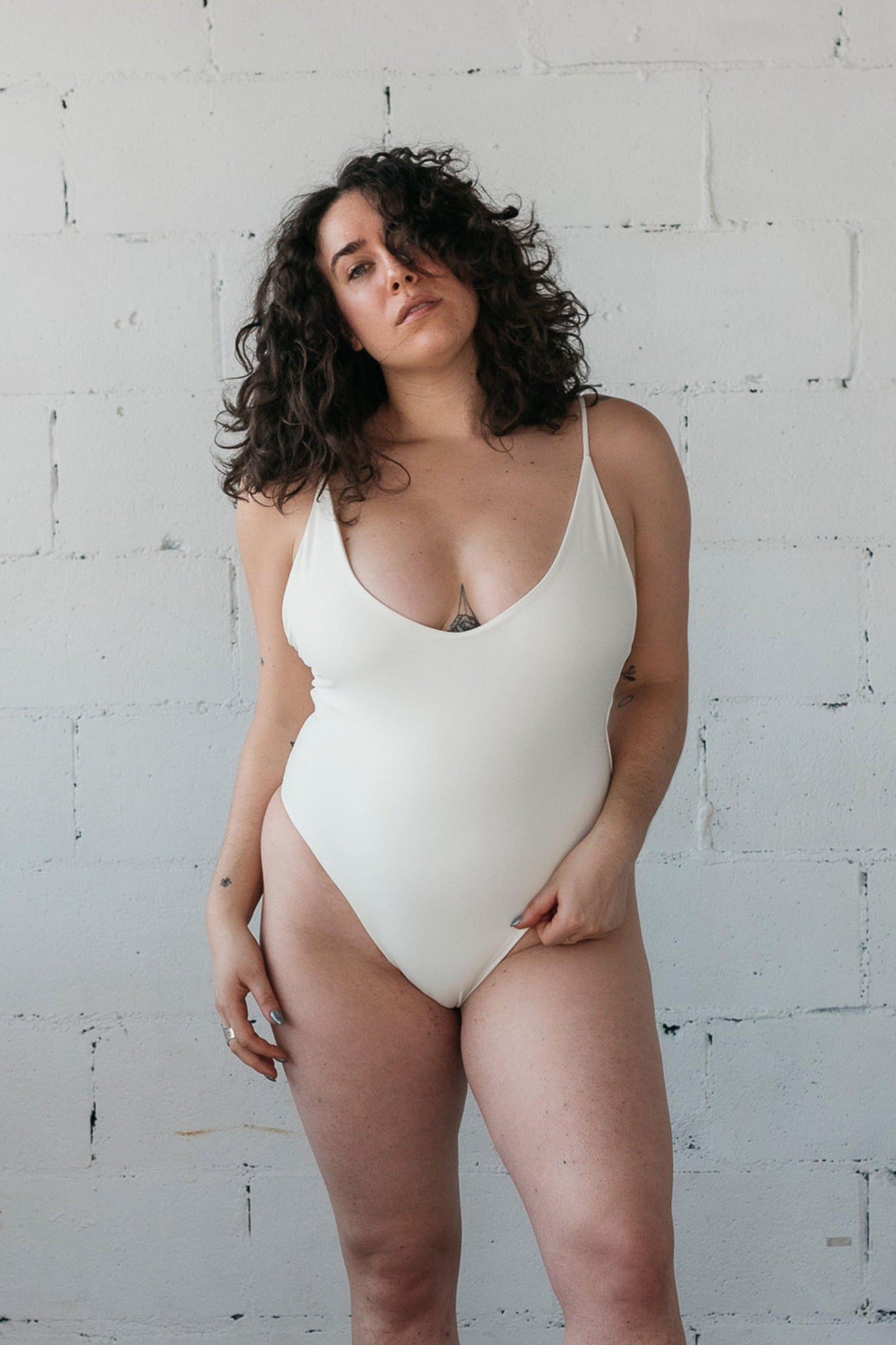 Woman standing in front of a white wall wearing a white one-piece swimsuit with spaghetti straps and a deep V in the front. 