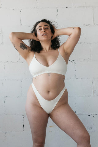 Woman standing against white wall wearing a white bikini featuring minimal coverage