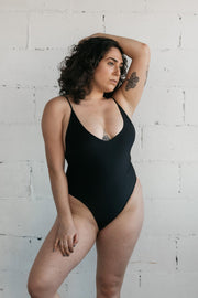 A woman standing against a white wall wearing a black one-piece swimsuit featuring spaghetti straps and a low neckline.