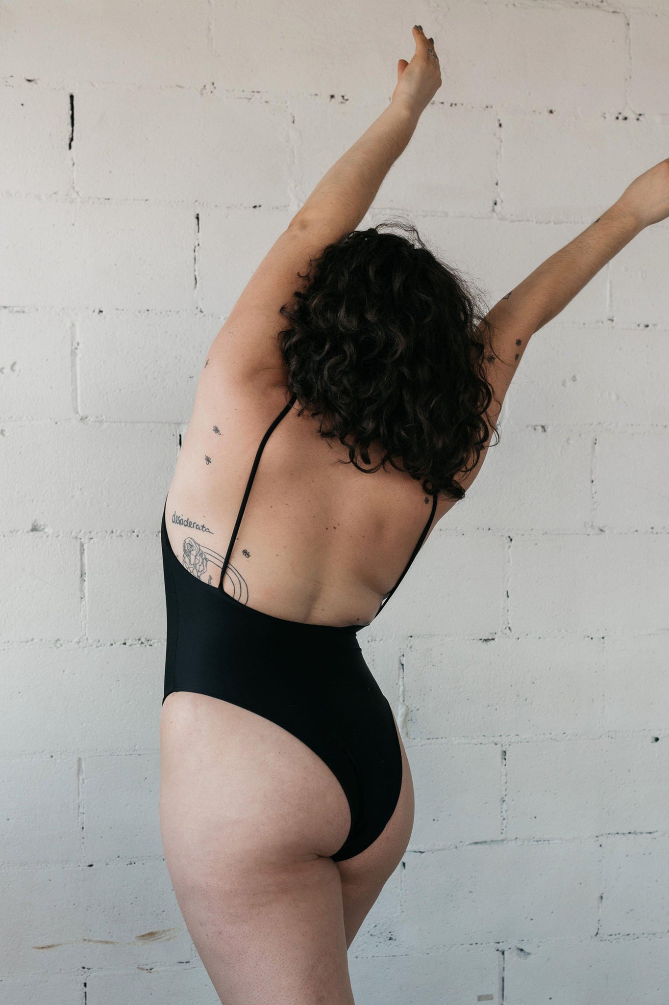 The back of a woman standing with her arms in the air wearing a black one-piece swimsuit featuring a low back and minimal coverage.