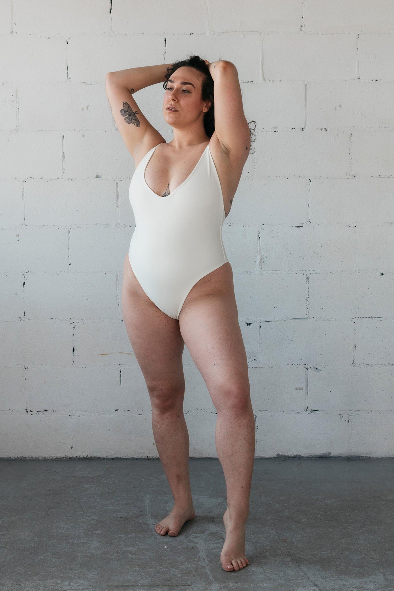 Woman standing with her hands on her head wearing a white one-piece swimsuit featuring a low cut neckline and spaghetti straps.