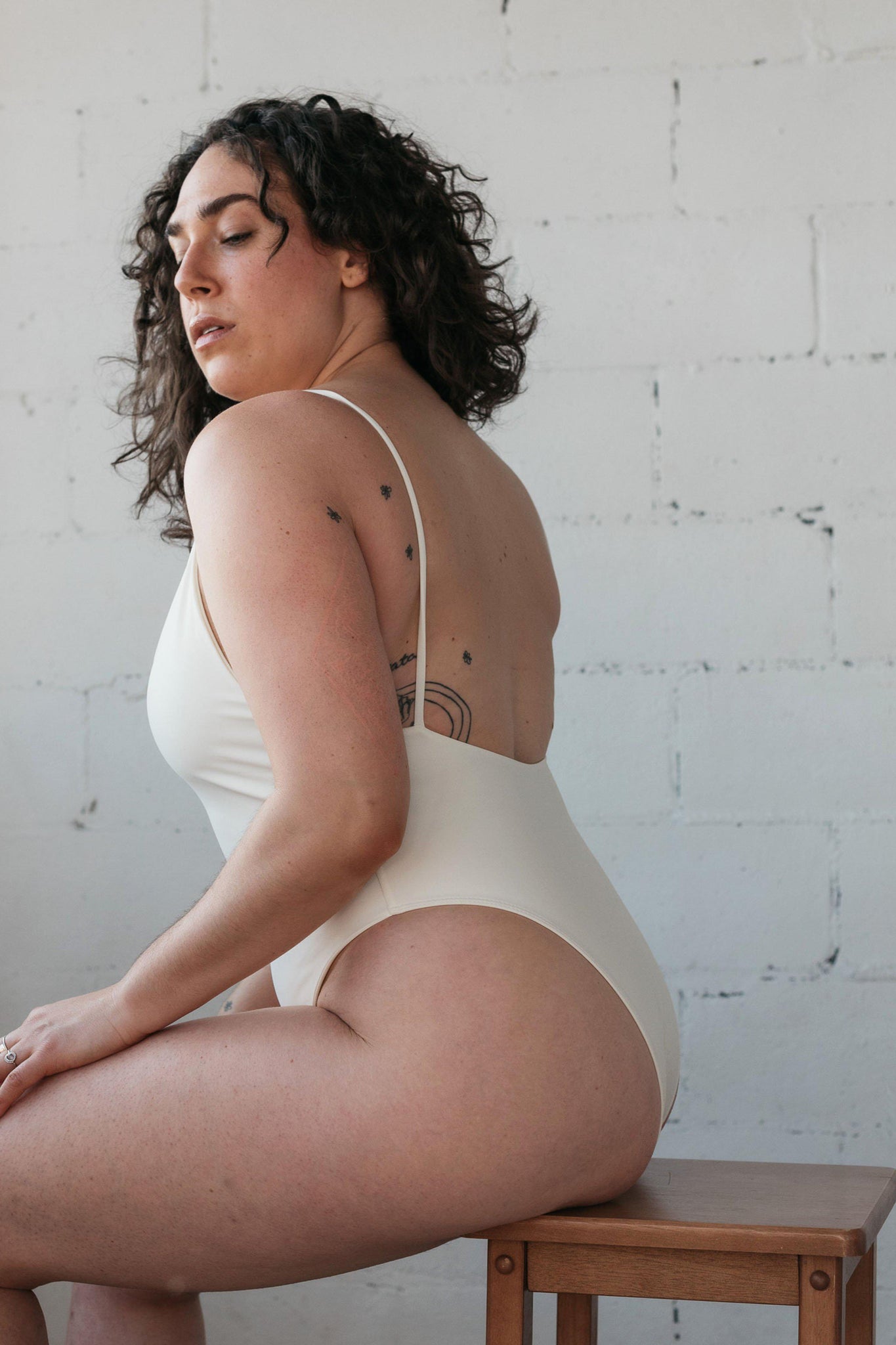 The side view of a woman sitting on a stool wearing a white one-piece swimsuit featuring spaghetti straps and a low back.