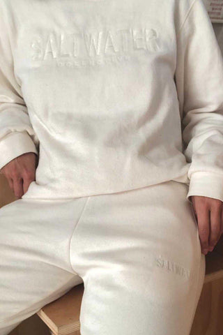 A woman sitting on a stool wearing a white oversized cotton crewneck and matching white cotton joggers with a tonal logo. 