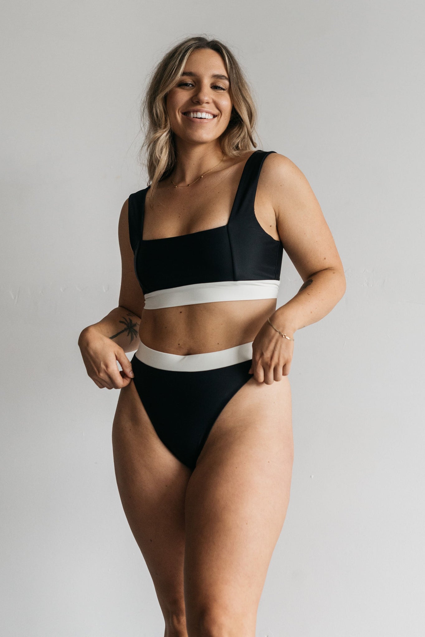 A woman standing in front of a white wall smiling wearing high waisted black bikini bottoms with a white waistband and a matching black bikini top with a square neckline.