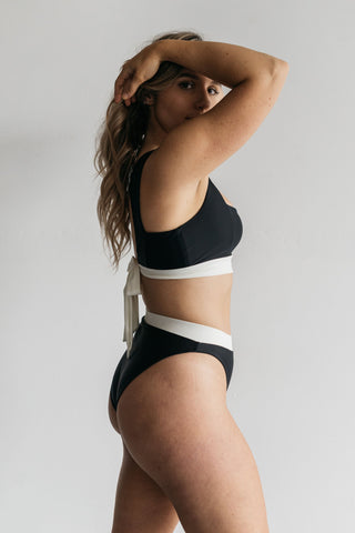 A woman standing to the side with her hands in her hair wearing high waisted black bikini bottoms with a white waistband and a matching black bikini top with an adjustable white tie.