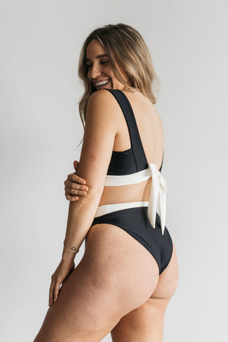 A woman smiling over her shoulder standing in front of a white wall wearing black and white high waisted bikini bottoms with a matching black and white bikini top.