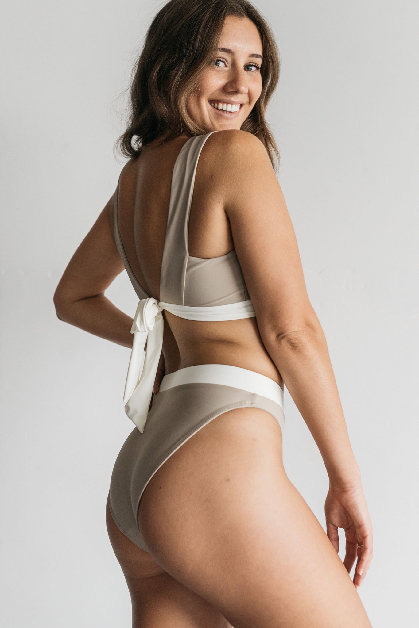 A woman smiling looking over her shoulder wearing beige bikini bottoms with a white waistband and a matching adjustable beige and white bikini top.