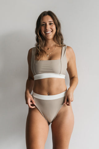 A woman standing with her hands on her hips wearing high waisted nude bikini bottoms with a white waistband and a matching nude bikini top with a white band.