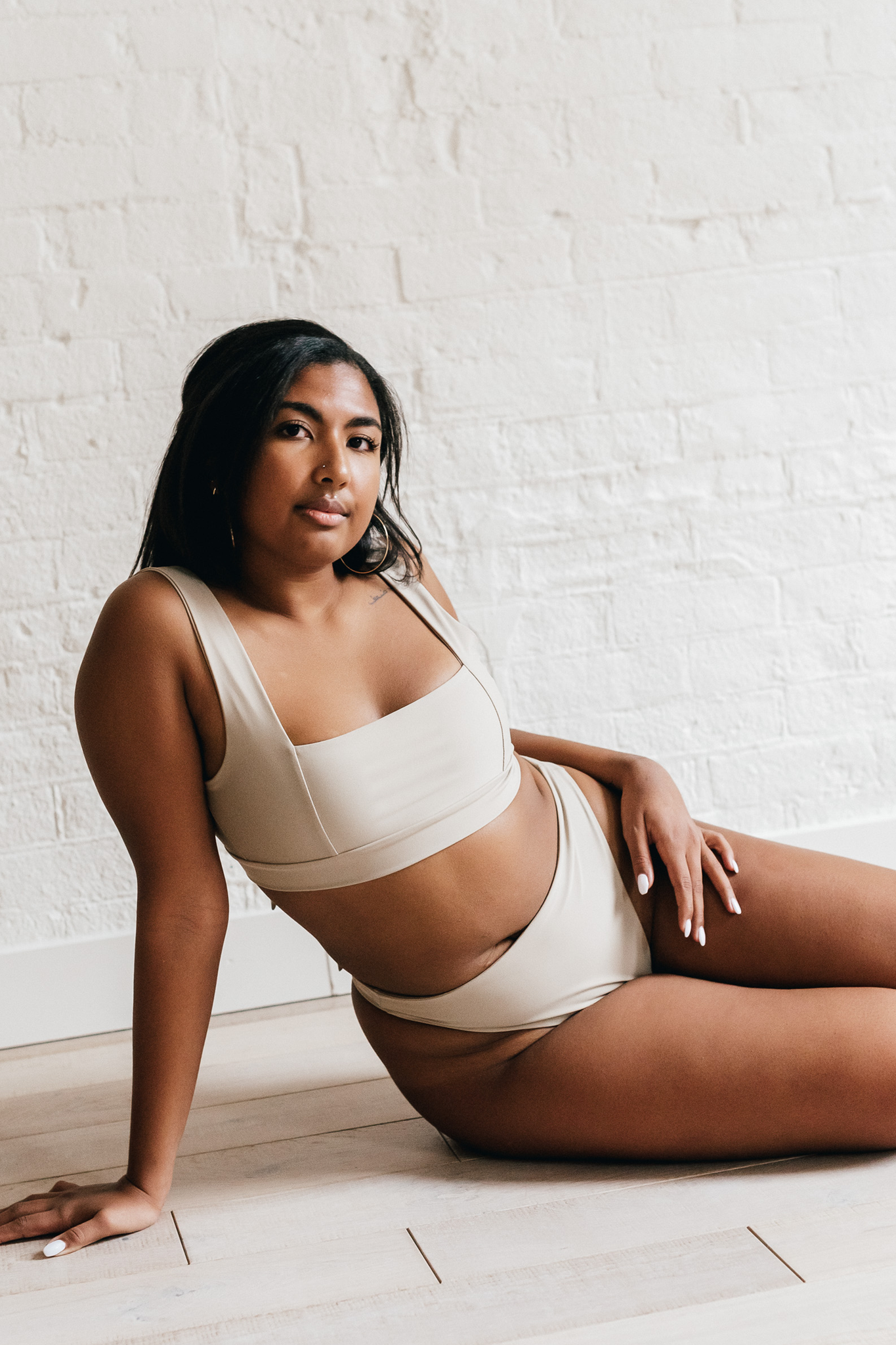 A woman leaning on one arm on the ground wearing beige high waisted bikini bottoms and a matching beige bikini top with a square neckline.