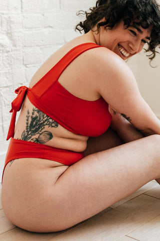 A woman sitting on the ground leaning over smiling  wearing bright red high cut bikini bottoms and a matching bright red bikini top with an adjustable tie.