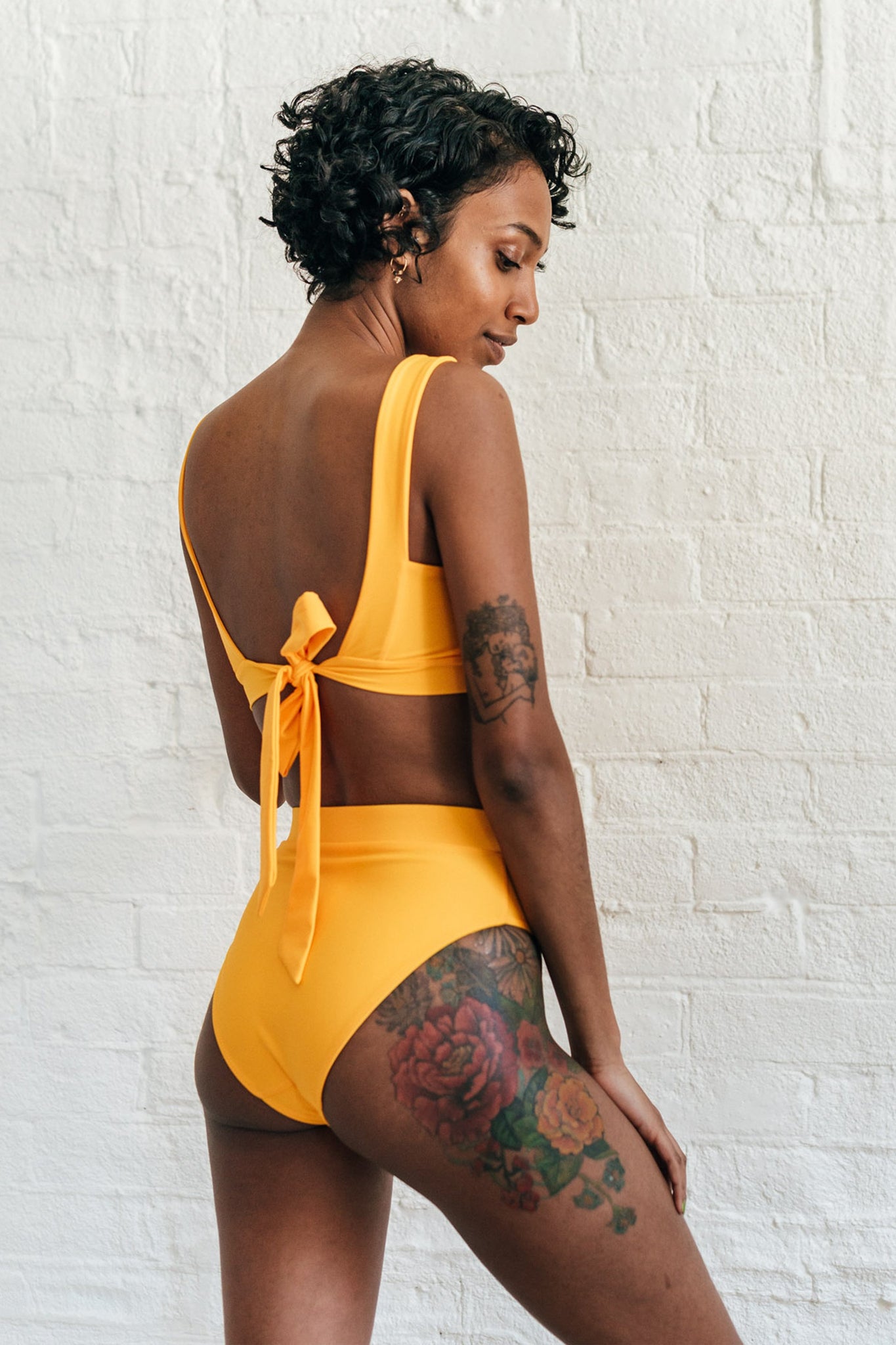 The back of a woman looking down wearing mustard yellow high waisted bikini bottoms and a matching mustard yellow adjustable bikini top.
