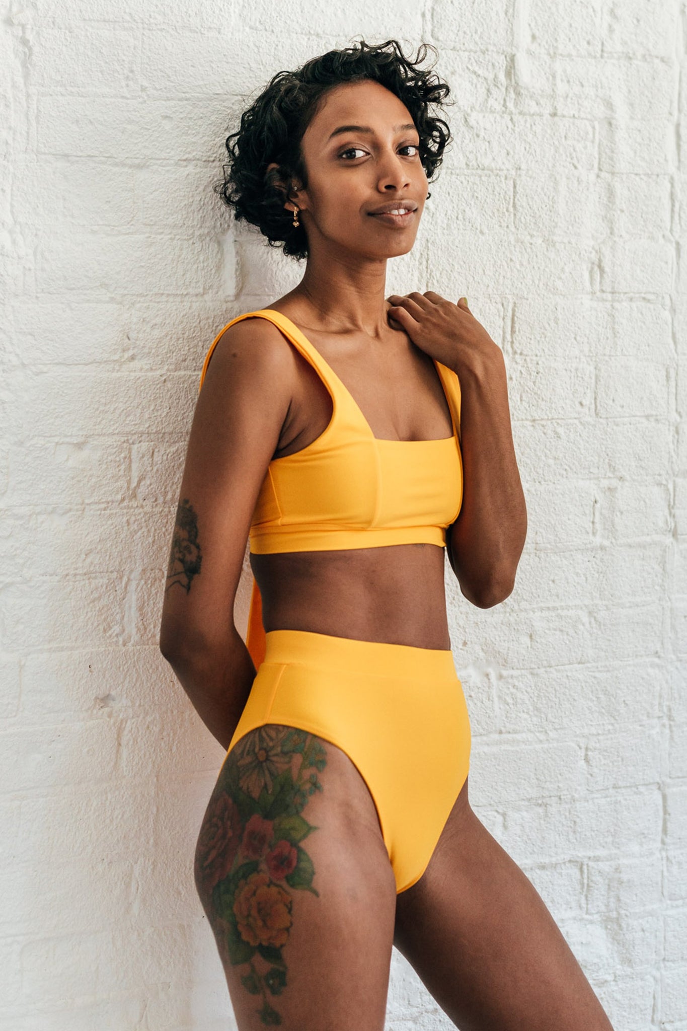 A woman leaning against a white brick wall with one hand on her shoulder wearing mustard yellow high waisted bikini bottoms and a matching mustard yellow bikini top with a square neckline. 
