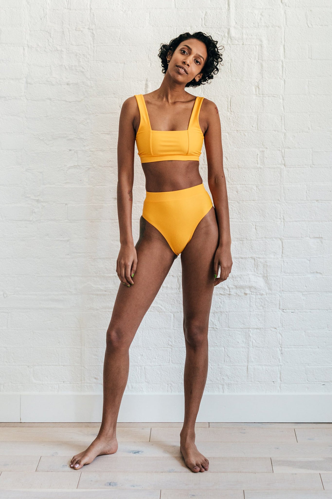 A woman standing in front of a white wall leaning her head to the side wearing mustard yellow high waisted bikini bottoms and a matching mustard yellow bikini top with a square neckline.