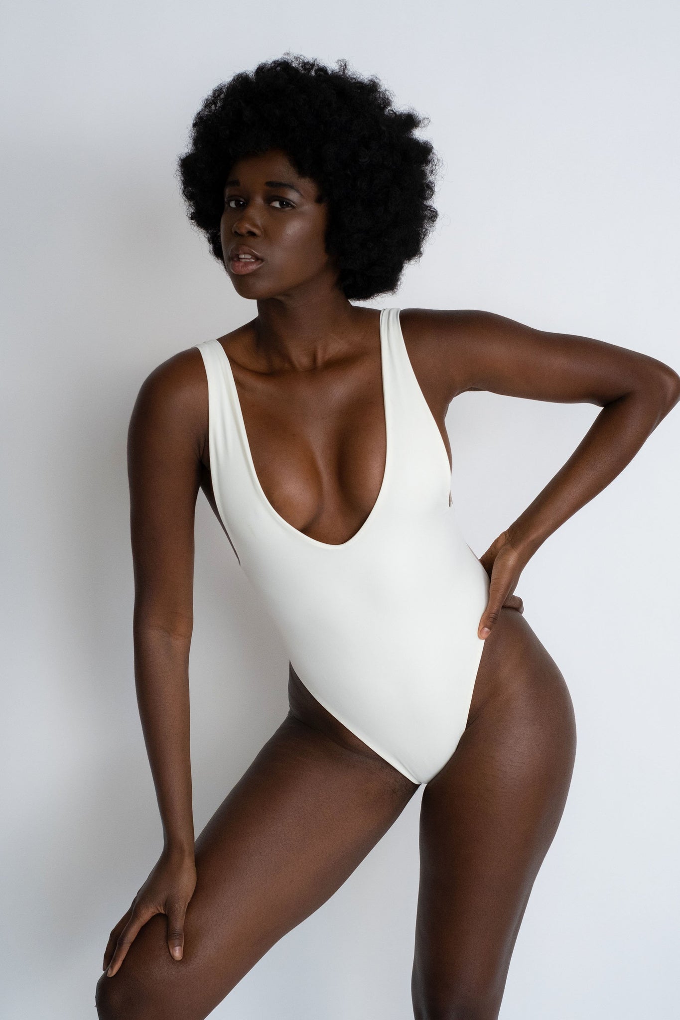 A woman standing with one hand on her hip wearing a white one piece swimsuit with a deep v neckline and minimal coverage.