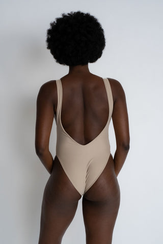 The back of a woman standing in front of a white wall wearing a nude one piece swimsuit with a low back and minimal coverage.