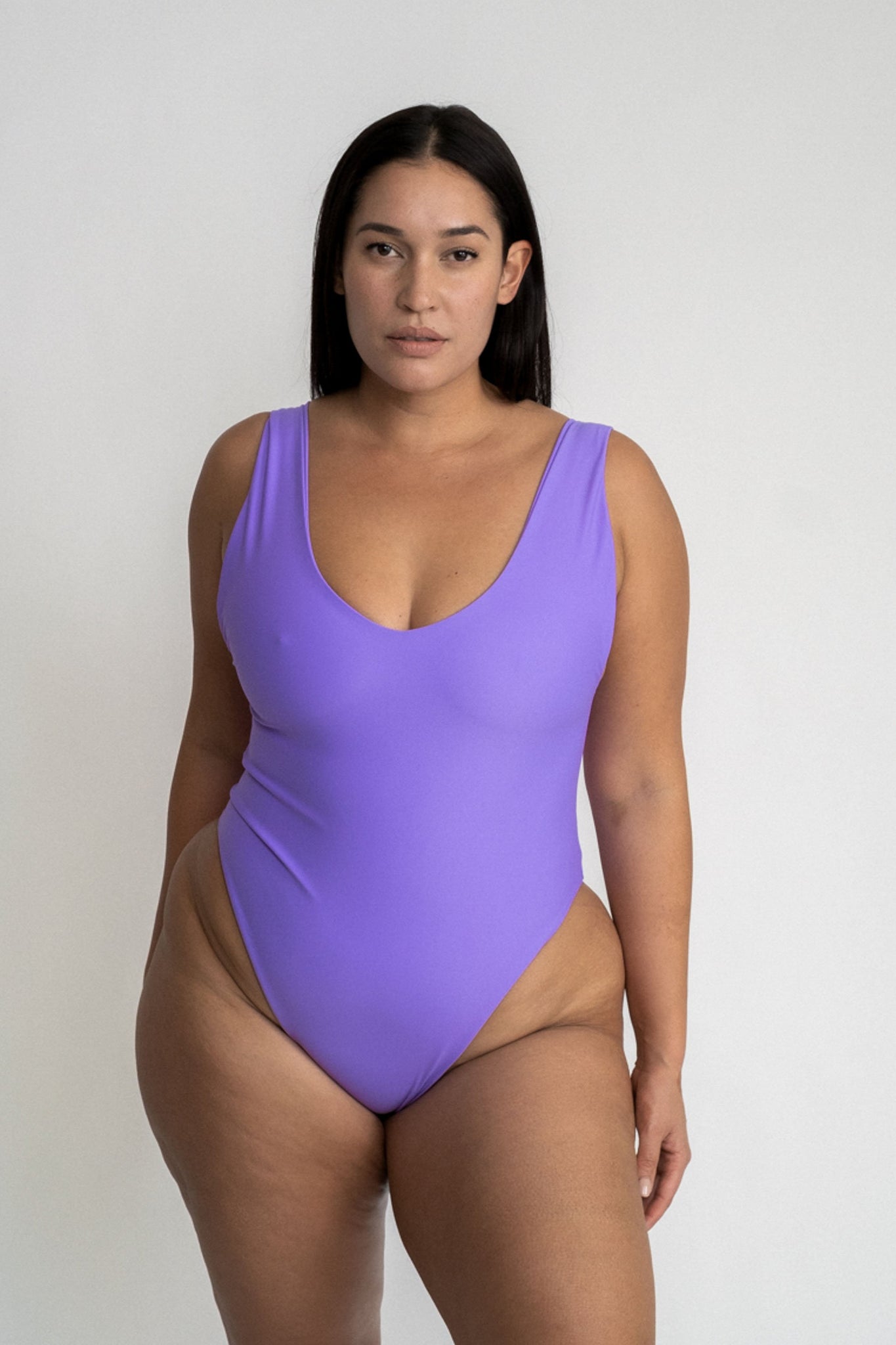 A woman standing in front of a white wall wearing a bright purple one piece swimsuit with a v neckline and minimal coverage.