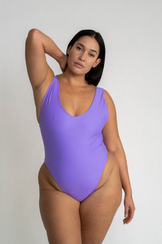 A woman standing with one hand in her hair wearing a bright purple one piece swimsuit with a v neckline and minimal coverage.