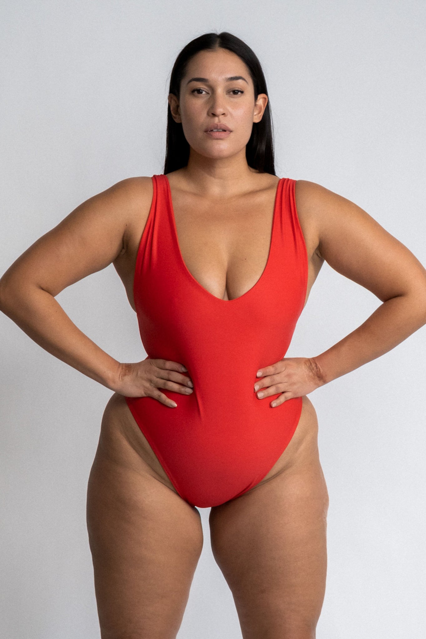 A woman standing in front of a white wall with her hands on her hips wearing a bright red one piece swimsuit with a v neckline and minimal coverage.