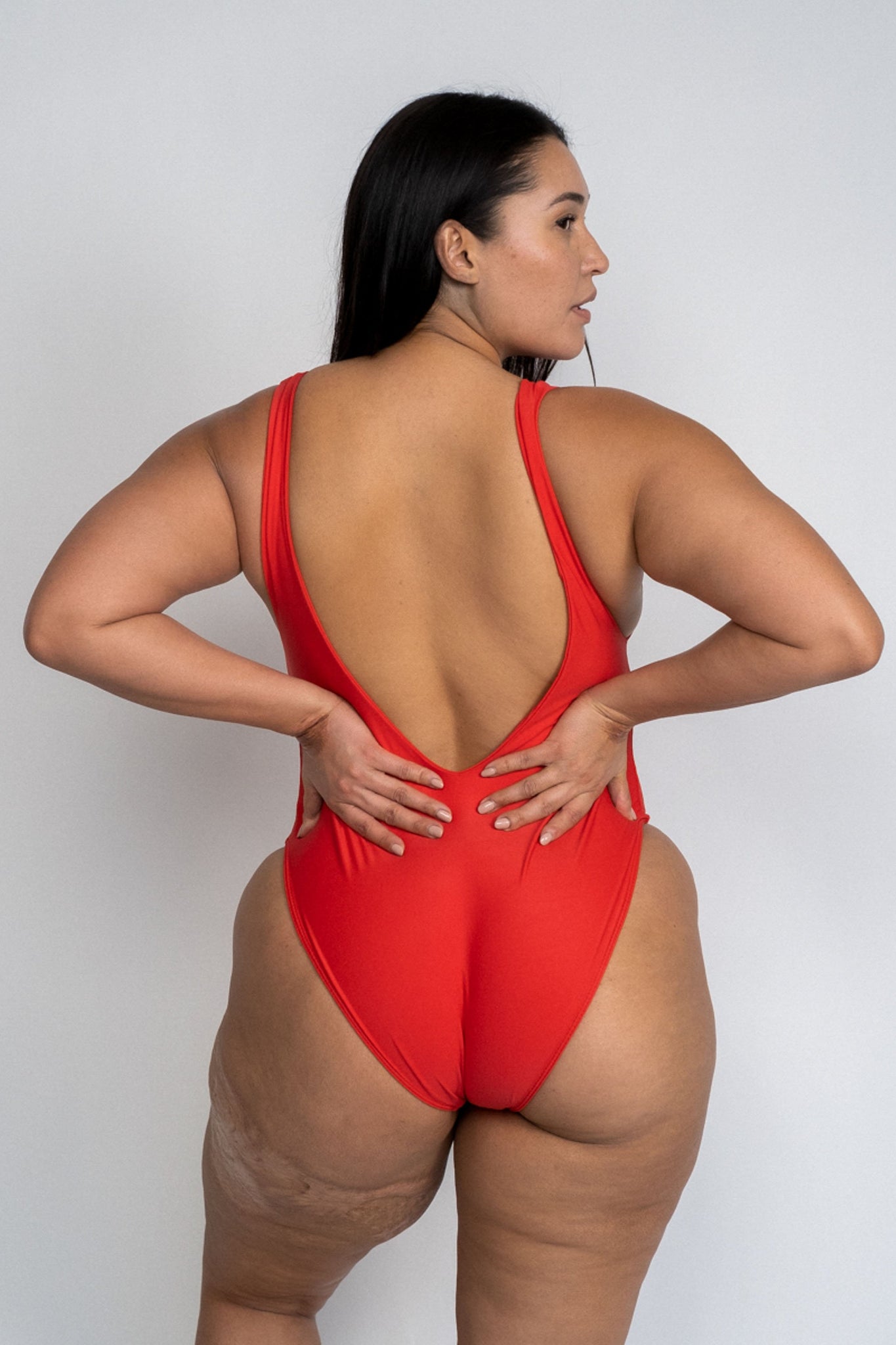 The back of a woman standing with her hands on her hips looking to the side wearing a bright red one piece swimsuit with a low back and minimal coverage.