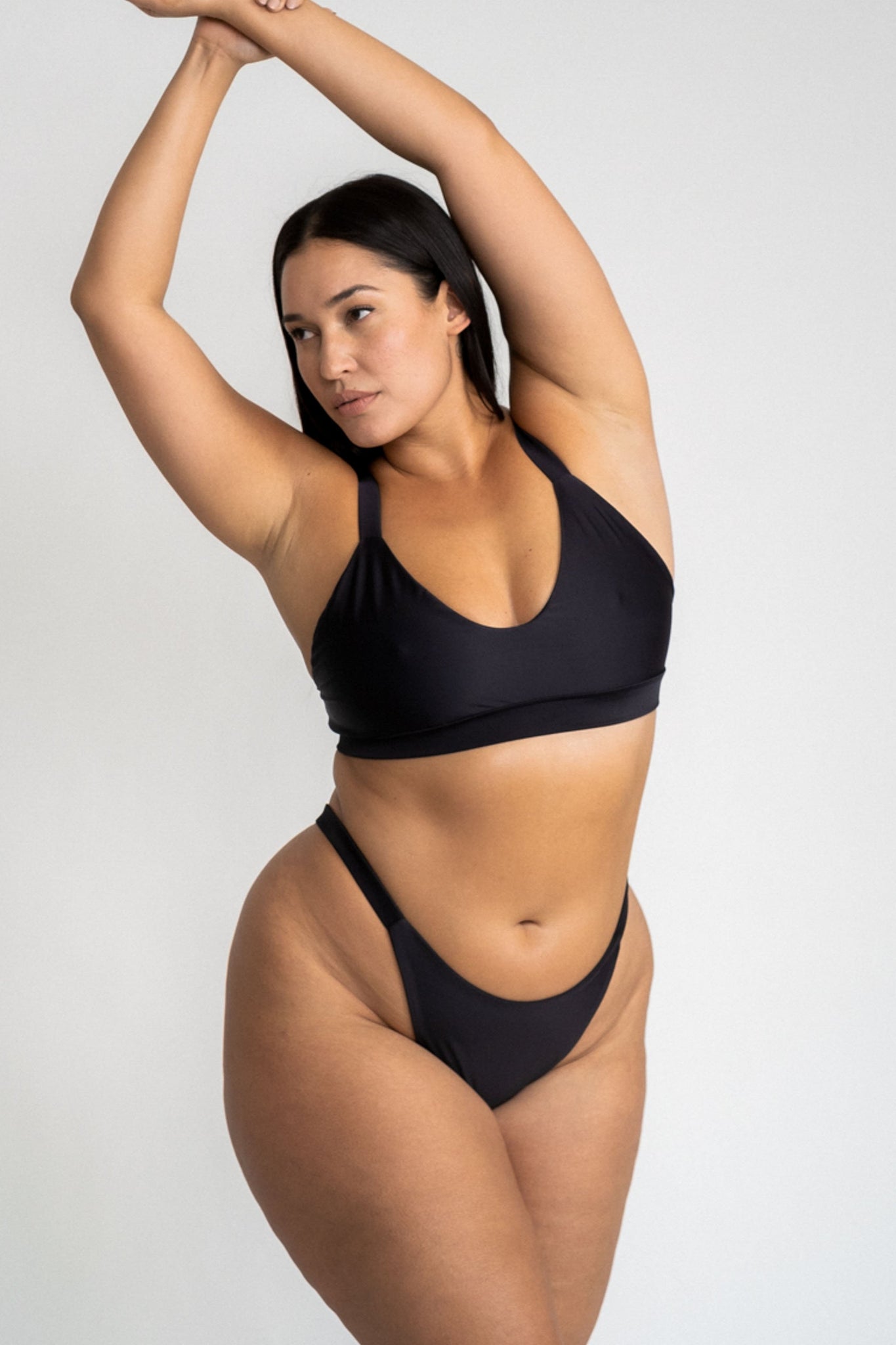 A woman standing with her arms above her head looking to the side wearing black high cut bikini bottoms and a matching black bikini top with a softly scooped neckline.