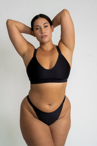 A woman standing in front of a white wall with her hands in her hair wearing black high cut bikini bottoms and a matching black bikini top with a softly scooped neckline.