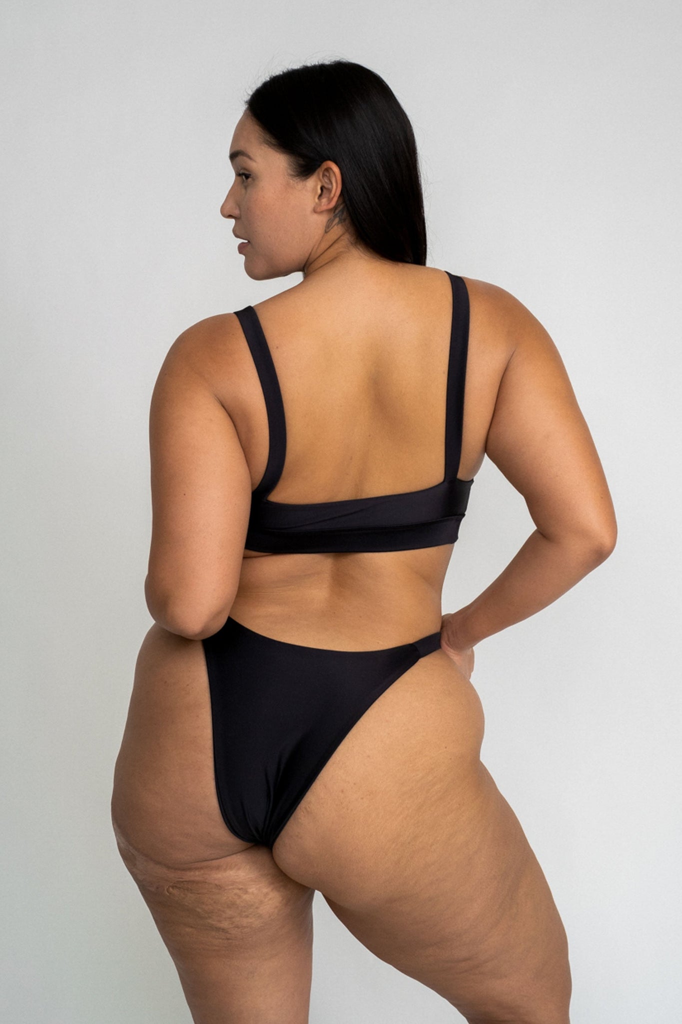 The back of a woman looking to the side wearing black high cut bikini bottoms and a matching black bikini top with thick supportive straps.