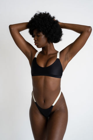 A woman standing with her hands in her hair looking down wearing black high cut bikini bottoms with white straps and a matching black bikini top with a softly scooped neckline and white supportive straps.