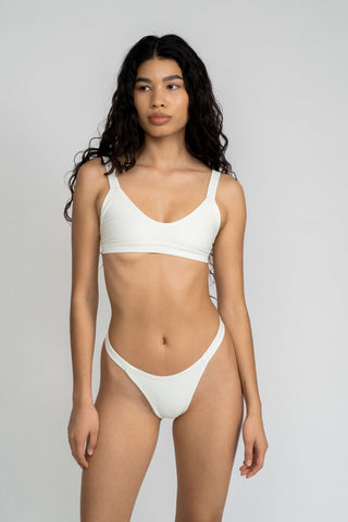 A woman standing in front of a white wall wearing white high cut bikini bottoms and a matching white bikini top with a softly scooped neckline.