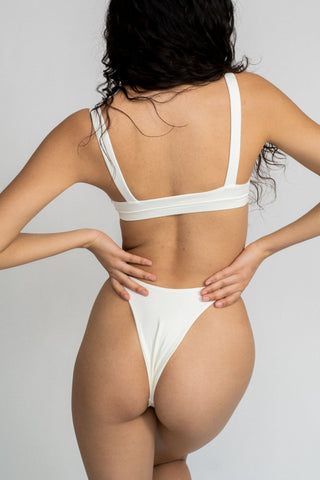 The back of a woman leaning to the side wearing white high cut bikini bottoms and a matching white bikini top with a softly scooped neckline.